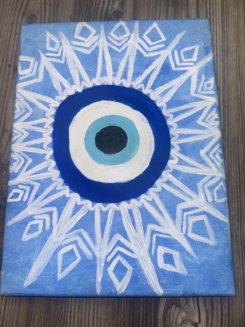 Puede incluir: Una pintura sobre lienzo azul y blanco con un dise&ntilde;o de mandala blanco que rodea un ojo turco azul. El ojo turco tiene un iris blanco y una pupila negra.