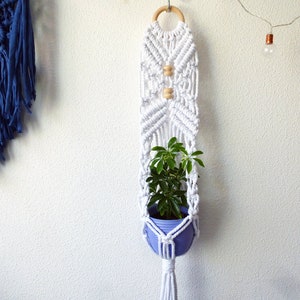 May include: White macrame plant hanger with a wooden ring at the top and two wooden beads. A small potted plant is hanging in the macrame.
