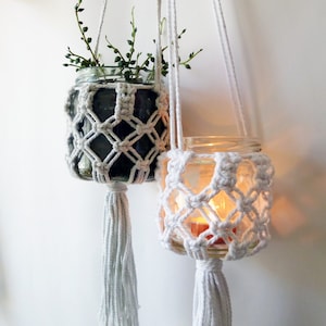 May include: Two white macrame plant hangers with glass jars. The top hanger has a small green plant inside the jar. The bottom hanger has a candle inside the jar.