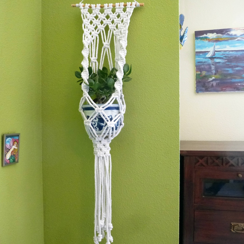 K&ouml;nnte beinhalten: Ein wei&szlig;er Makramee-Pflanzenh&auml;nger mit einer kleinen Topfpflanze, die daran h&auml;ngt. Der H&auml;nger ist mit einer Holzleiste an einer Wand befestigt.