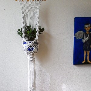 K&ouml;nnte beinhalten: Ein wei&szlig;er Makramee-Pflanzenh&auml;nger mit einer kleinen Topfpflanze, die in der Mitte h&auml;ngt. Der Pflanzenh&auml;nger hat eine lange Quaste unten.