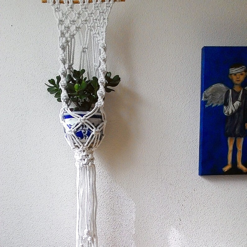 K&ouml;nnte beinhalten: Ein wei&szlig;er Makramee-Pflanzenh&auml;nger mit einer kleinen Topfpflanze, die in der Mitte h&auml;ngt. Der Pflanzenh&auml;nger hat eine lange Quaste unten.