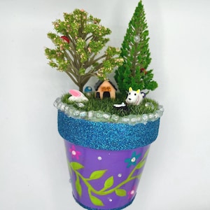 May include: A whimsical miniature garden in a purple flower pot. The pot has a blue glitter band and is decorated with painted flowers and vines. Inside, a tiny house, trees, and animal figurines create a charming scene.