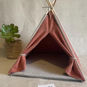 May include: A small, coral-colored pet teepee with a gray interior and trim. The tent is supported by wooden poles tied together at the top. A small succulent plant in a woven pot sits to the left. The label "Pöticha Pet Shop" is visible.