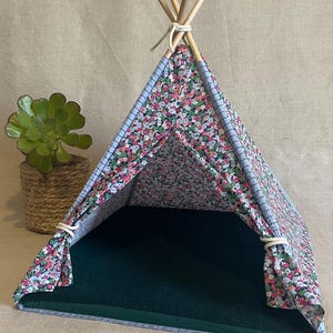 Könnte beinhalten: Ein Haustier-Tipi mit einem Außenmaterial aus floralem Stoff und einem dunkelgrünen Innenraum. Das Tipi wird oben von Holzpfählen getragen und hat eine hellblaue Umrandung. Links vom Tipi steht eine Topfpflanze. Der Text "Pâticha Pet Shop" ist sichtbar.