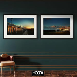 Peut inclure: Deux tirages encadrés de paysages. L'un montre des cabines de plage au coucher du soleil, l'autre un restaurant au crépuscule. Le ciel passe du bleu à l'orange. Les cadres sont noirs et le mur est vert foncé.