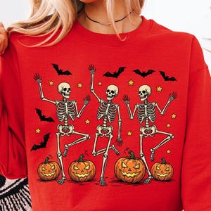 May include: A red Halloween sweatshirt featuring three dancing skeletons, jack-o'-lanterns, bats, and stars. The long-sleeved crew neck top is a festive garment for the spooky season.