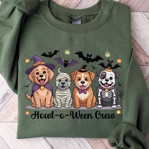 May include: An olive green sweatshirt featuring a Halloween-themed graphic. The graphic depicts four cartoon dogs dressed in Halloween costumes, including a witch, mummy, and skeleton. The text "Howl-o-Ween Crew" is below the dogs, with bats and pumpkins above.