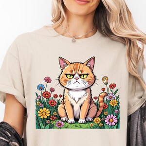 May include: A beige t-shirt with a cartoon image of an orange tabby cat sitting in a field of flowers. The cat has a grumpy expression.