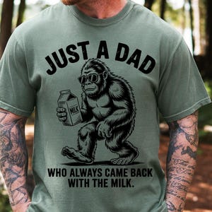 May include: Olive green t-shirt with the text "JUST A DAD" above a cartoon gorilla wearing sunglasses and holding a milk carton. The text "WHO ALWAYS CAME BACK WITH THE MILK" is below the gorilla.