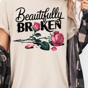 May include: Beige t-shirt with the words "Beautifully Broken" in black script and block letters. A watercolor-style rose with pink petals and green leaves is below the text. The person is wearing a black and gray plaid shirt.