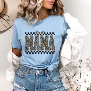 May include: Light blue t-shirt with the word "MAMA" in a camouflage design, framed by a black and white checkered pattern. A small bow is above the word. The shirt is paired with denim shorts.