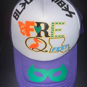 May include: A white trucker hat with a purple brim. The front of the hat has a colourful design with the words "FRE" and "FEST" and a green heart. The back of the hat has the words "BLUU" and "VIBX". A green masquerade mask is on the brim of the hat.