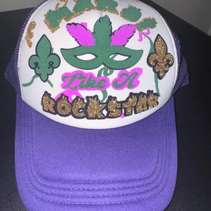 Op de afbeelding: Een witte trucker-pet met een paarse mesh-rug. De voorkant van de pet heeft gouden glitterletters die "Mardi Gras Like A Rockstar" zeggen en een groen en roze Mardi Gras-masker.