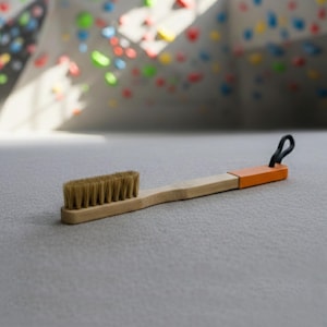 Op de afbeelding: Een bamboe klimborstel met natuurlijke borstelharen en een oranje handvat. De borstel heeft een zwarte lus om op te hangen. De achtergrond toont een klimmuur met kleurrijke grepen.