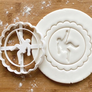 Könnte beinhalten: Ein weißer Kunststoff-Ausstecher mit gewelltem Rand, der eine Turnerin in einer dynamischen Pose zeigt. Daneben ein Teigkreis mit dem Abdruck der Turnerin. Die Szene befindet sich auf einer mit Mehl bestäubten Holzoberfläche.