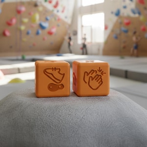 Pode incluir: Dois cubos laranja com ícones relacionados à escalada. Um cubo apresenta um sapato e uma pegada, o outro mostra duas mãos aplaudindo. Os cubos estão sobre uma superfície cinza, com um ginásio de escalada ao fundo.