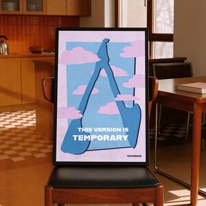 Può includere: Una stampa artistica incorniciata con una figura astratta blu su sfondo azzurro, con nuvole rosa. Il testo "THIS VERSION IS TEMPORARY" è in basso. La stampa è esposta su una sedia di legno.