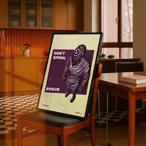 May include: A framed poster with the words "DON'T SPIRAL EVOLVE" and an illustration of a figure with zebra-like stripes. The poster is propped on a wooden chair with a black seat, in a room with a kitchen and a table.
