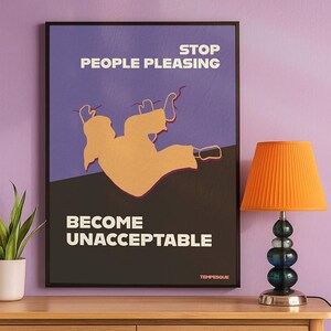 May include: A framed poster with the text "STOP PEOPLE PLEASING" and "BECOME UNACCEPTABLE" in white. The poster features a gold-colored illustration of a person falling. An orange lamp with a pleated shade sits on a wooden surface.