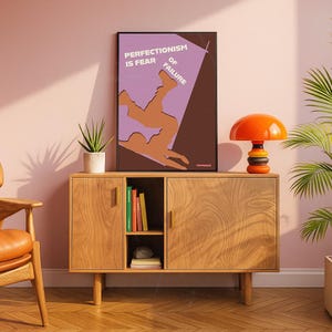 K&ouml;nnte beinhalten: Ein Holzschrank mit einem gerahmten Poster mit der Aufschrift "Perfektionismus ist Angst vor dem Scheitern". Auf dem Schrank steht eine orangefarbene und schwarze Lampe. Links vom Schrank steht eine Topfpflanze.