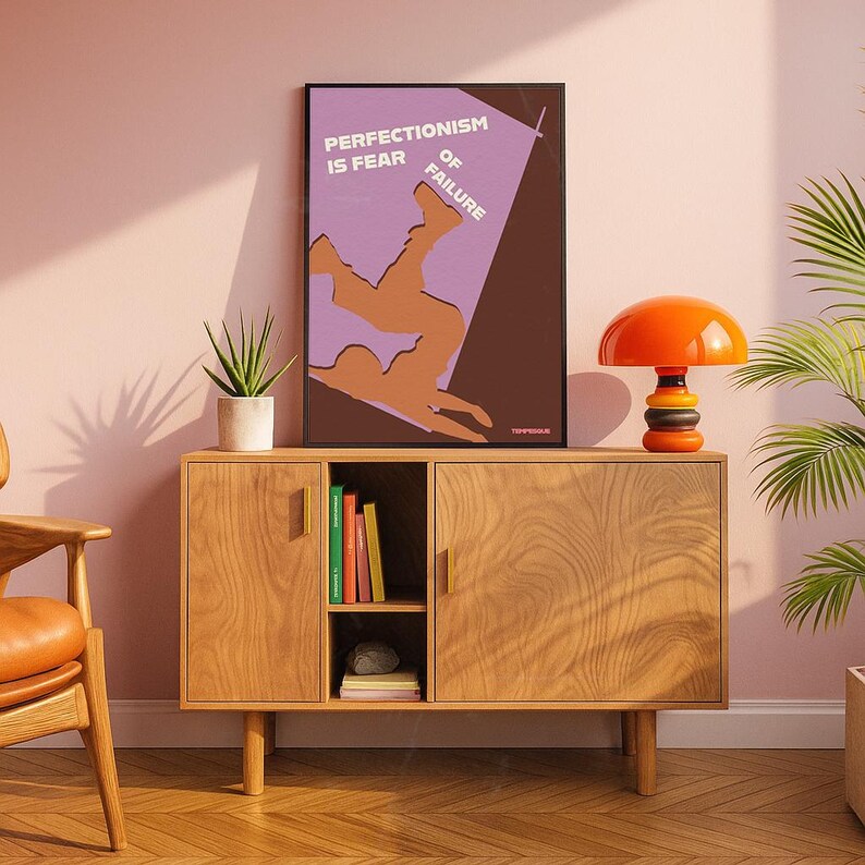 K&ouml;nnte beinhalten: Ein Holzschrank mit einem gerahmten Poster mit der Aufschrift "Perfektionismus ist Angst vor dem Scheitern". Auf dem Schrank steht eine orangefarbene und schwarze Lampe. Links vom Schrank steht eine Topfpflanze.