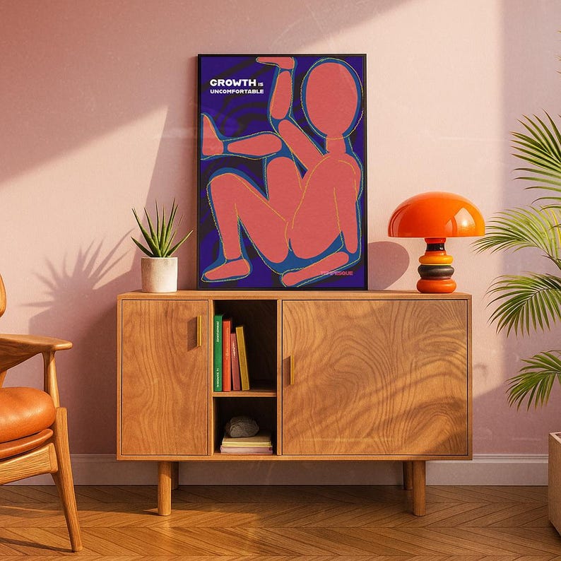 May include: A framed art print with the words "GROWTH. UNCOMFORTABLE" above a red figure on a blue background. The print is displayed on a wooden cabinet with an orange lamp and a potted plant. The room has a mid-century modern aesthetic.