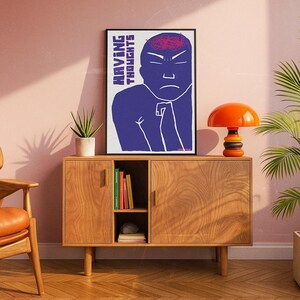 May include: A framed art print with the text "HAVING THOUGHTS" and a blue figure with a red brain. The print is displayed above a wooden cabinet with a small orange lamp and a potted plant. The cabinet has two doors and a shelf with books.