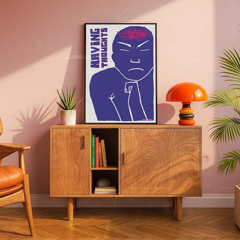 May include: A framed art print with the text "HAVING THOUGHTS" and a blue figure with a red brain. The print is displayed above a wooden cabinet with a small orange lamp and a potted plant. The cabinet has two doors and a shelf with books.
