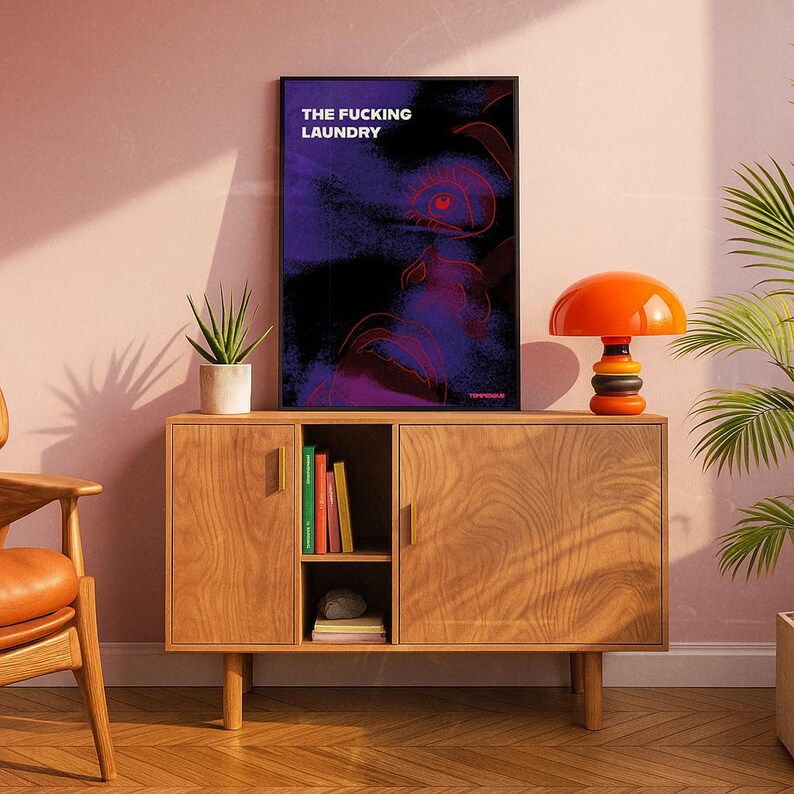 Op de afbeelding: Een ingelijste kunstdruk met de tekst "THE FUCKING LAUNDRY" in het wit. Het kunstwerk heeft een donkere achtergrond met paarse en rode abstracte vormen. Een oranje paddenstoelvormige lamp staat op een houten kast.