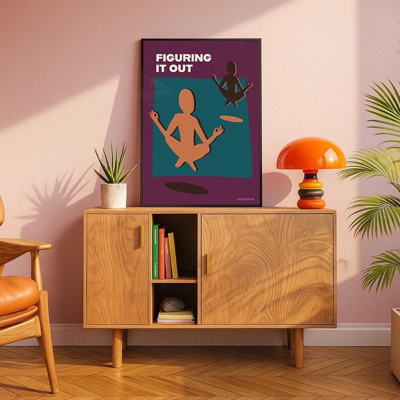 May include: A framed art print with the text "FIGURING IT OUT" features a meditating figure in a yoga pose. The artwork is displayed on a wooden cabinet with an orange lamp and a potted plant. The cabinet has two doors and a shelf with books.