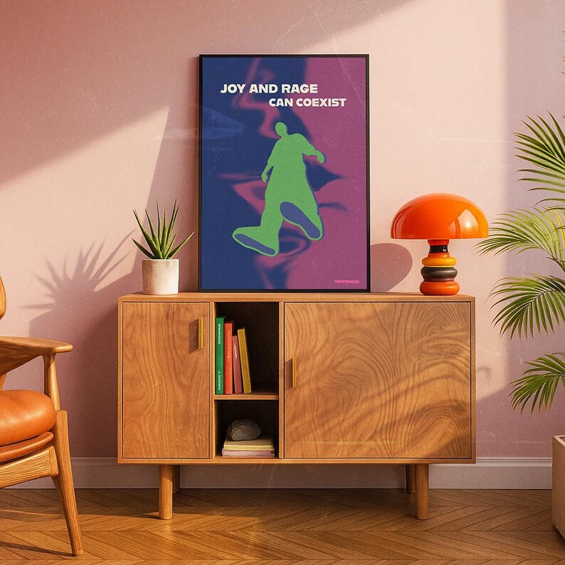 May include: A framed art print with the text "JOY AND RAGE CAN COEXIST" and a green figure on a blue and purple background. The print is displayed on a wooden cabinet with an orange lamp and a potted plant.