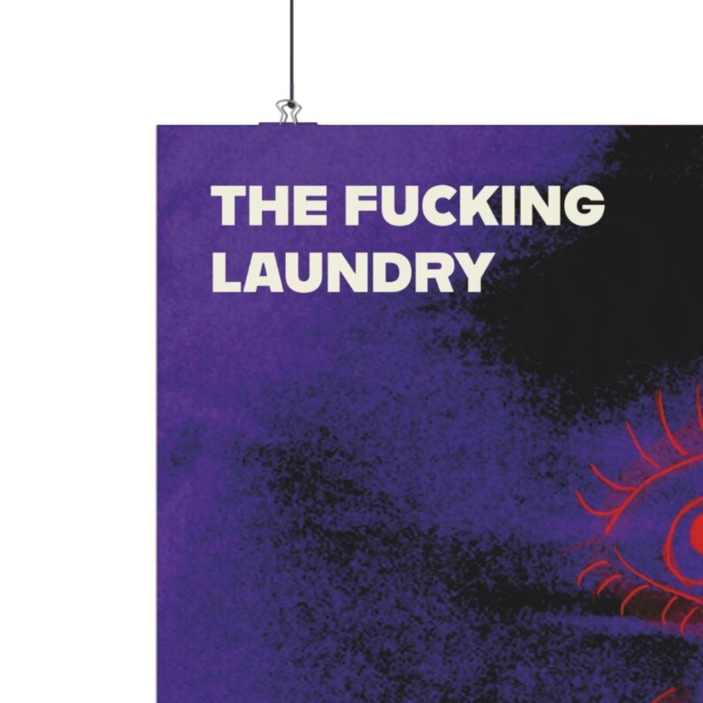 Op de afbeelding: Een paarse poster met de tekst "THE FUCKING LAUNDRY" in het wit. De poster wordt opgehangen door een zilveren clip aan een zwart koord. Een rode en zwarte oogafbeelding is zichtbaar in de rechter benedenhoek.