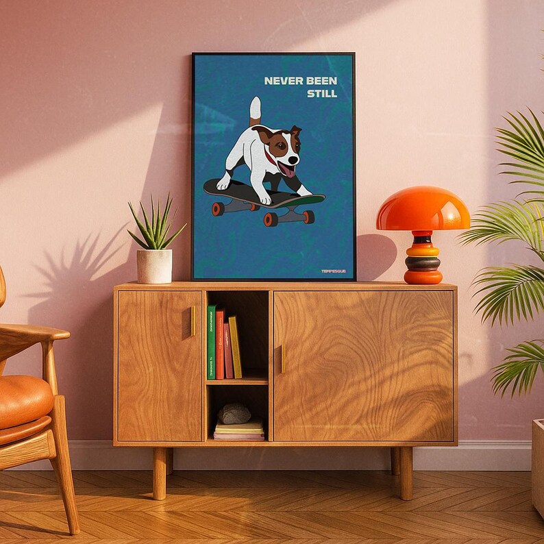 May include: A framed art print featuring a dog on a skateboard with the text "NEVER BEEN STILL" on a blue background. The artwork is displayed on a wooden cabinet with an orange lamp and a potted plant.