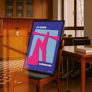 Può includere: Una stampa artistica incorniciata con le parole "I'M DONE SHRINKING" su sfondo blu. L'opera presenta una figura astratta rosa con le braccia tese su un rettangolo azzurro. La stampa è esposta su una sedia di legno.
