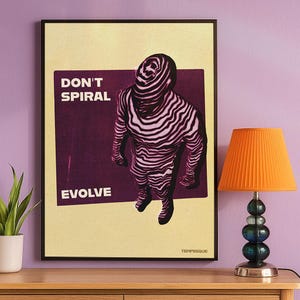 May include: Framed art print featuring a figure with a zebra-like pattern, set against a purple backdrop. The words "DON'T SPIRAL EVOLVE" are displayed in white. The print is displayed on a wooden surface, next to a lamp with an orange lampshade.