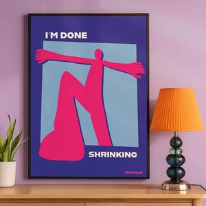 May include: A framed art print with the words "I'M DONE SHRINKING" in white text. The artwork features a pink figure with outstretched arms against a light blue square on a dark blue background. An orange lamp is to the right.