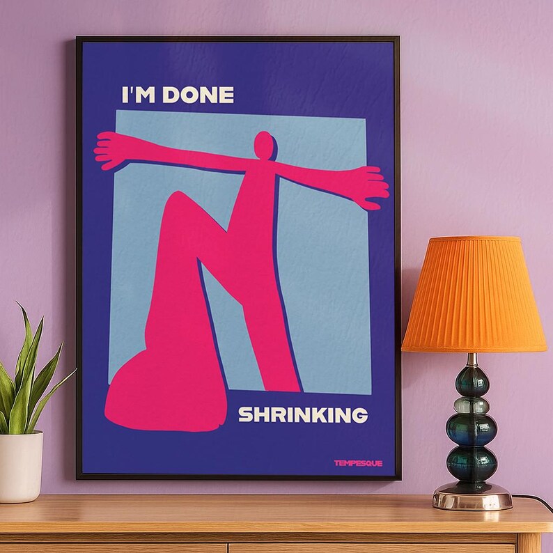 May include: A framed art print with the words "I'M DONE SHRINKING" in white text. The artwork features a pink figure with outstretched arms against a light blue square on a dark blue background. An orange lamp is to the right.