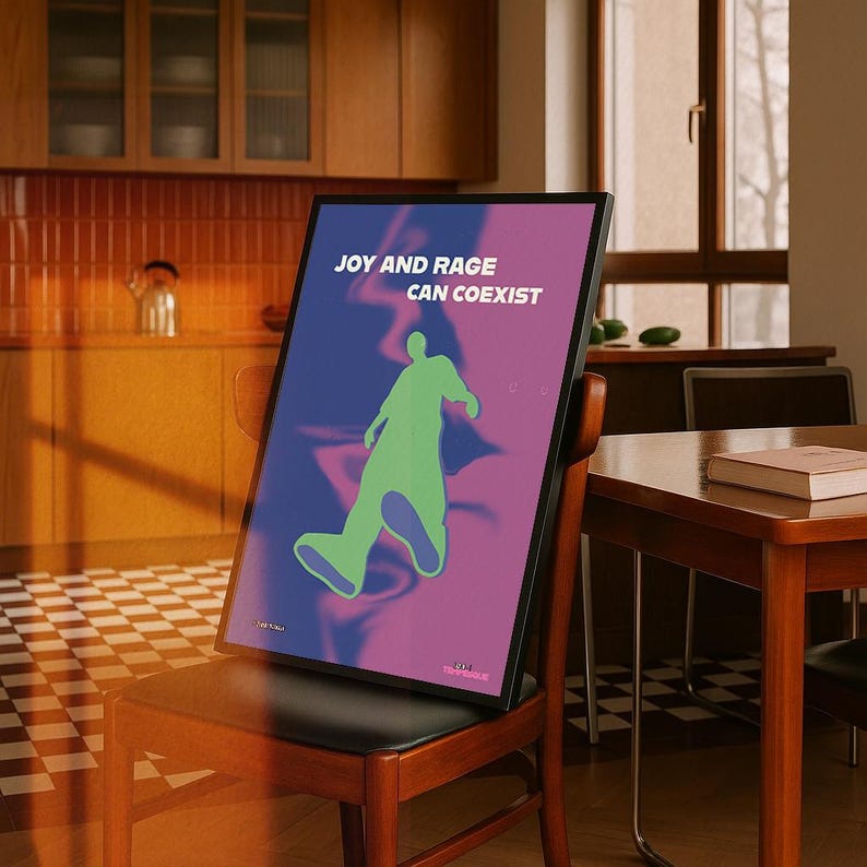 May include: A framed poster with the text "JOY AND RAGE CAN COEXIST" in white. The poster features a green silhouette of a person walking on a gradient background of blue and purple. The poster is propped on a wooden chair.