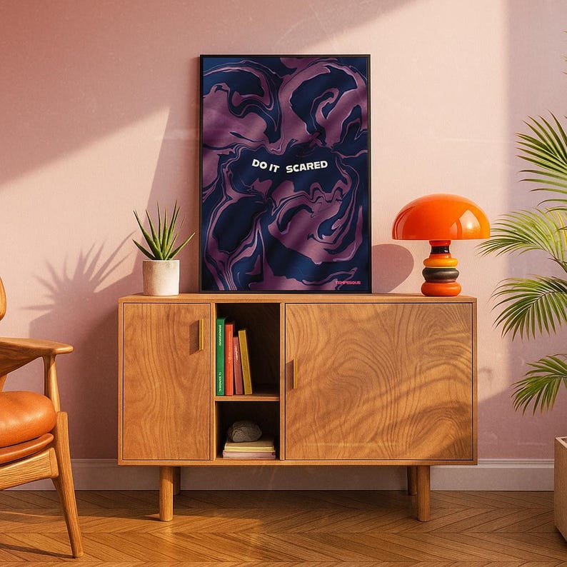 May include: A wooden cabinet with a framed art print and a mushroom-shaped lamp. The art print features a swirling purple and navy design with the words "DO IT SCARED". The lamp is orange and black. A potted plant sits on top of the cabinet.