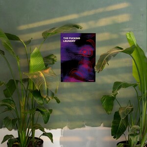 Op de afbeelding: Een poster met de tekst "THE FUCKING LAUNDRY" in het wit, tegen een donkerpaars en zwart abstract ontwerp met rode accenten. De poster hangt aan een groene muur, geflankeerd door grote potplanten.