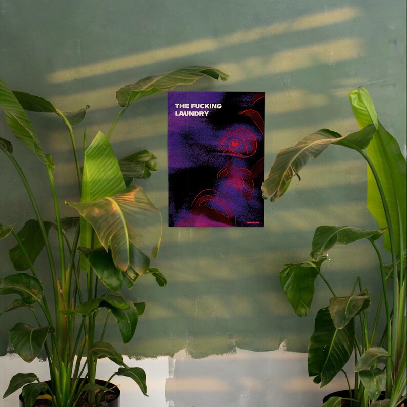 Op de afbeelding: Een poster met de tekst "THE FUCKING LAUNDRY" in het wit, tegen een donkerpaars en zwart abstract ontwerp met rode accenten. De poster hangt aan een groene muur, geflankeerd door grote potplanten.