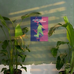 May include: A poster with the text "JOY AND RAGE CAN COEXIST" and a green silhouette of a person walking. The background transitions from blue to purple. The poster is mounted on a green wall, with large green plants in the foreground.