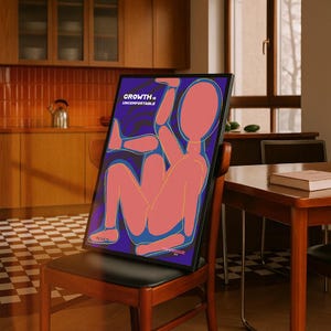 May include: A framed art print featuring a stylized human figure in shades of pink and blue against a dark purple background. The text "GROWTH is UNCOMFORTABLE" is displayed at the top. The artwork is propped on a wooden chair.