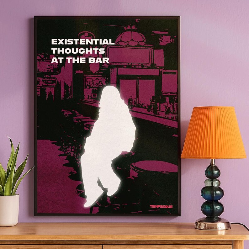Op de afbeelding: Een ingelijste kunstdruk met de tekst "EXISTENTIAL THOUGHTS AT THE BAR" in het wit. De print toont een silhouet van een persoon in een baromgeving met een paars en zwart kleurenschema. Een oranje lamp met een geplooide kap staat op een houten oppervlak.