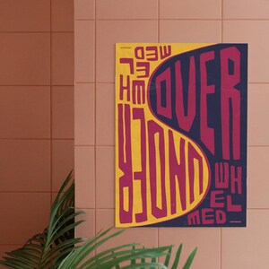 Può includere: Un poster con le parole "OVERWHELMED UNDER" in un carattere audace e stilizzato. Il testo è principalmente magenta e giallo su uno sfondo blu scuro. Il poster è appeso a una parete piastrellata rosa.