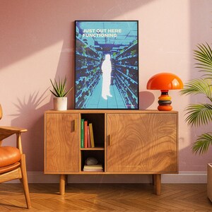 K&ouml;nnte beinhalten: Ein Holzschrank mit einem gerahmten Poster mit der Aufschrift "JUST OUT HERE FUNCTIONING". Das Poster zeigt eine Person in einem Lebensmittelgesch&auml;ft. Auf dem Schrank steht eine orangefarbene und schwarze Lampe. Links vom Schrank steht eine Topfpflanze.