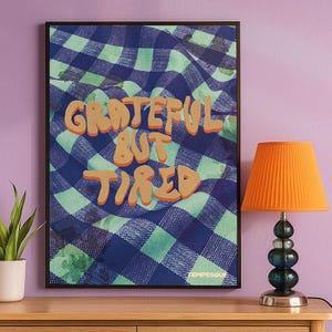 Può includere: Una stampa artistica incorniciata con le parole "Grateful But Tired" in lettere arancioni su un motivo a scacchi blu e verde. Una lampada arancione con paralume plissettato è su una superficie di legno.