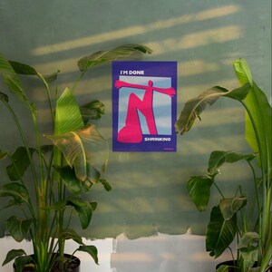 May include: A poster with the words "I'M DONE" and "SHRINKING" in white text. The poster features a pink figure with outstretched arms against a blue and white background. The poster is mounted on a green wall, with green plants in the foreground.