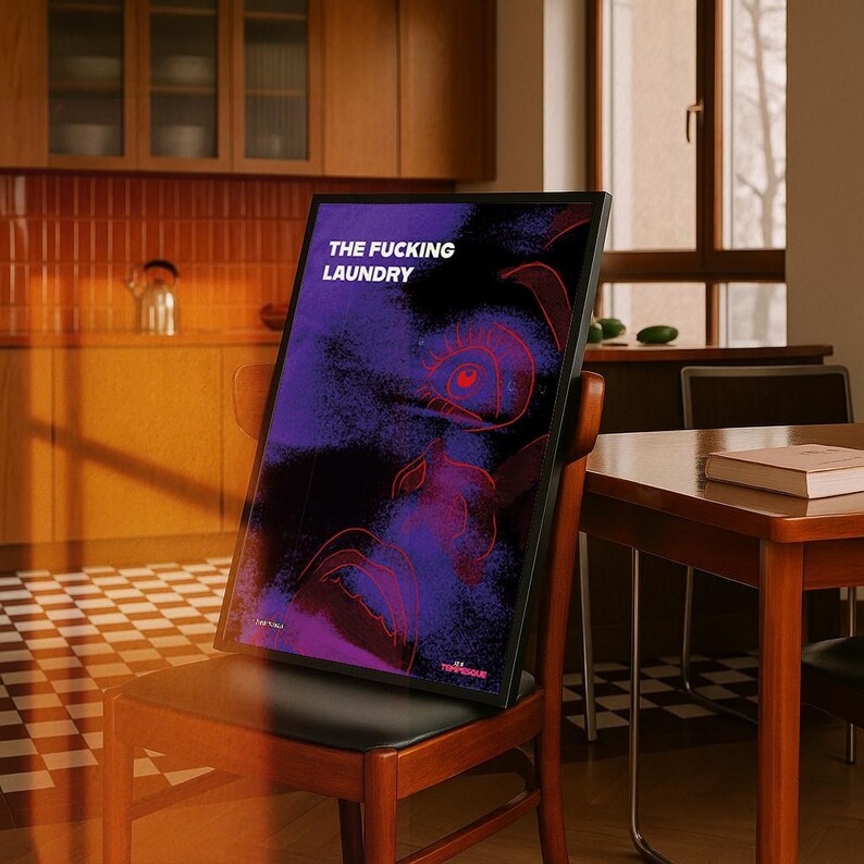 Op de afbeelding: Een ingelijste kunstdruk met de tekst "THE FUCKING LAUNDRY" in het wit. Het kunstwerk heeft een paars en zwart abstract ontwerp met rode lijnen. De print staat op een houten stoel in een keuken.